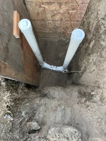 PVC pipes installed in a trench for plumbing, surrounded by dirt and wood support.